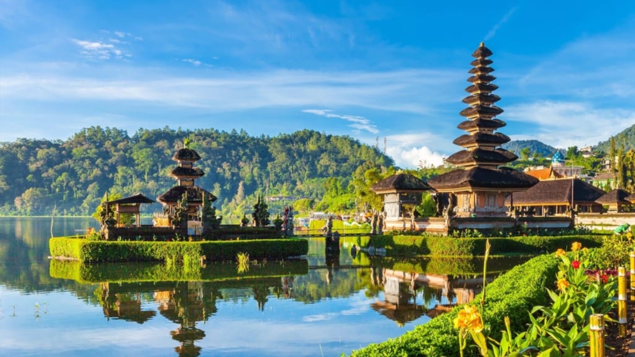 "Scenic view of the Ulun Danu Beratan Temple in Bali, Indonesia, situated on the shores of Lake Bratan. The temple's pagoda-style shrines and structures are reflected in the calm lake waters, with lush green hills and a clear blue sky in the background."