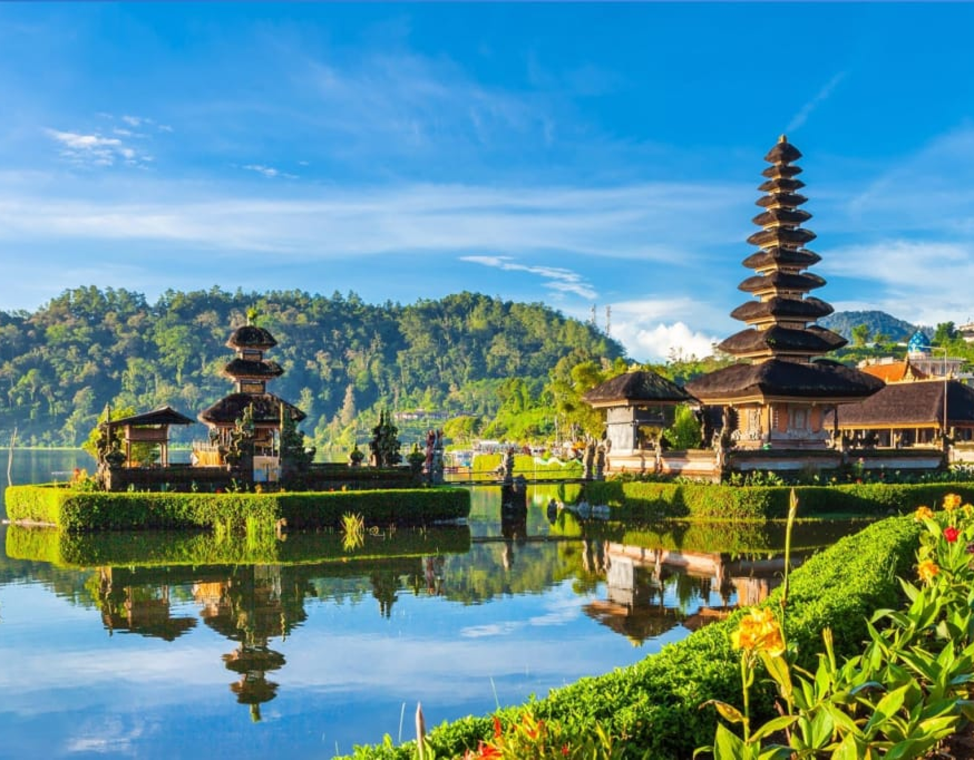 "Scenic view of the Ulun Danu Beratan Temple in Bali, Indonesia, situated on the shores of Lake Bratan. The temple's pagoda-style shrines and structures are reflected in the calm lake waters, with lush green hills and a clear blue sky in the background."