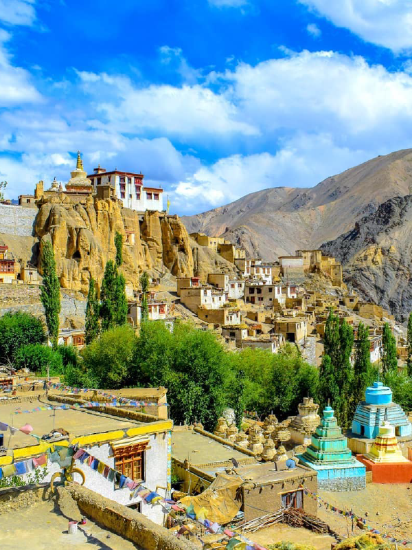 "A scenic view of the verdant fields and scattered houses in the valley of Leh-Ladakh, with barren mountains rising in the background under a partly cloudy sky."